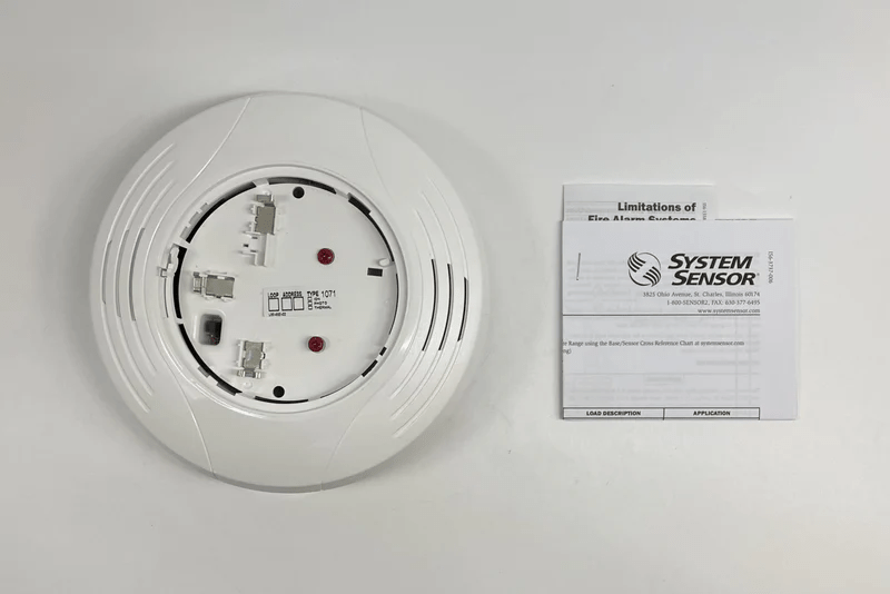 System Sensor B224RB-WH fire alarm detector with white housing, mounted on a ceiling in a building.